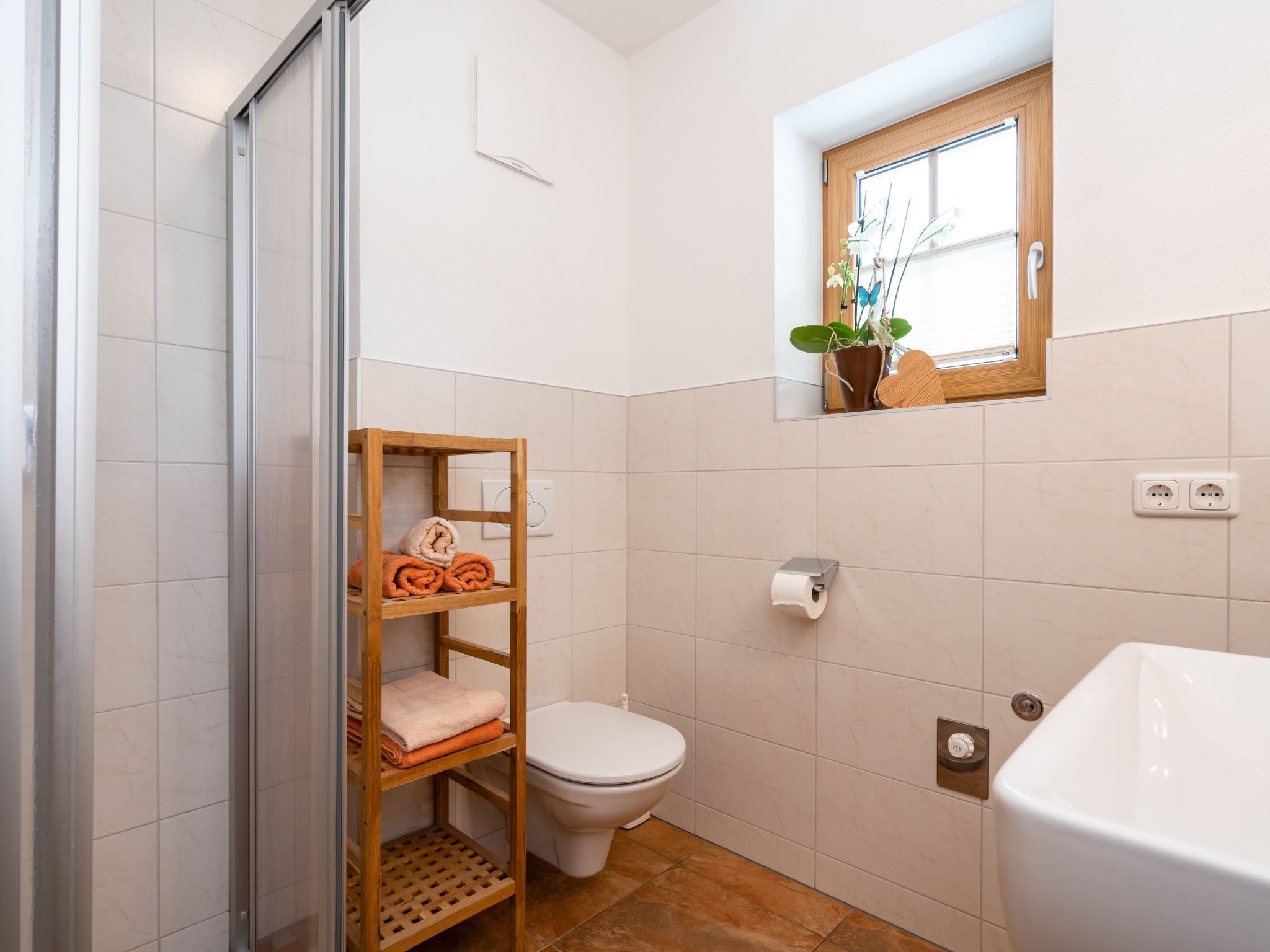 A bathroom with a toilet, a wooden shelf with towels, a window, and a sink.
