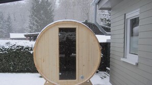 Una sauna in legno con una porta, situata su un pontile coperto di neve di fronte a una casa.