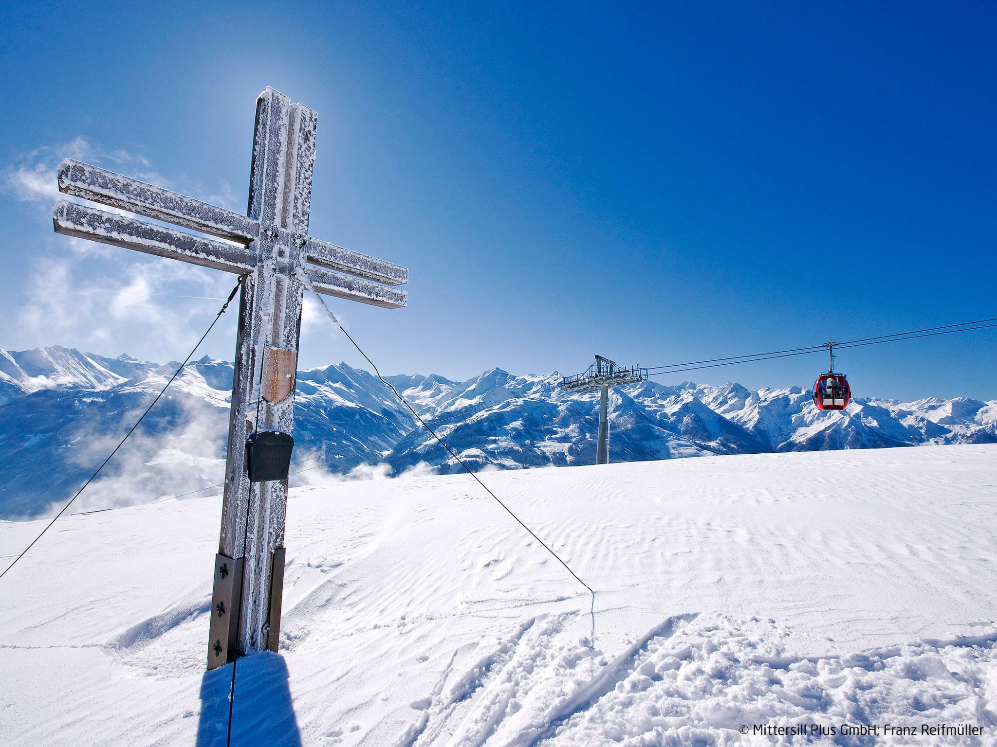 A snow-covered mountain with a wooden cross, a cable car, and a clear blue sky.