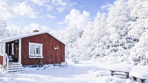 En röd trästuga med en veranda och trappsteg som leder upp till entrén, omgiven av snötäckta träd.