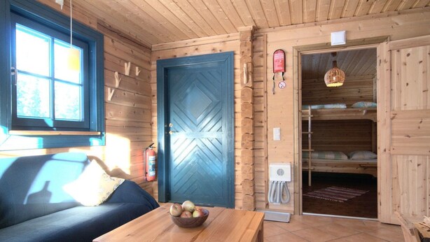 Un interior de cabaña acogedor con techo de madera, una puerta azul, un sofá, una mesa con manzanas, literas y una lámpara colgante.