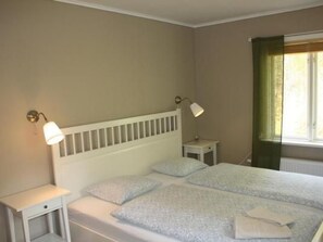 Ein Schlafzimmer mit einem weißen Kopfteil, zwei Nachttischen, einem Bett mit einem gemusterten Bettbezug und einem Fenster mit grünen Vorhängen.