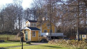 Une maison jaune avec une petite dépendance attenante, entourée d’arbres et d’une zone herbeuse.
