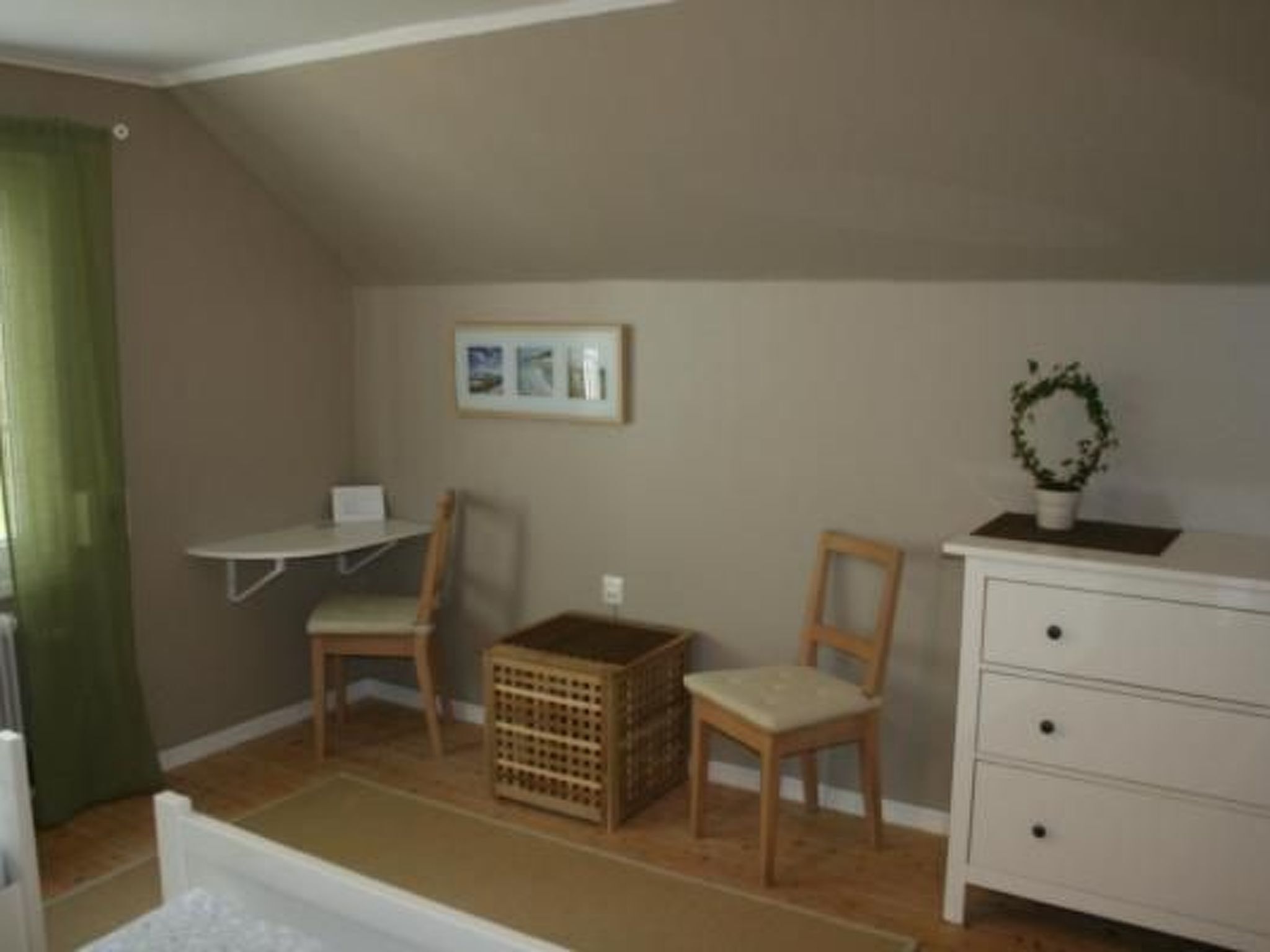 A small, cozy room with a sloped ceiling, a wooden chair, a white dresser, a small table, and a framed picture on the wall.