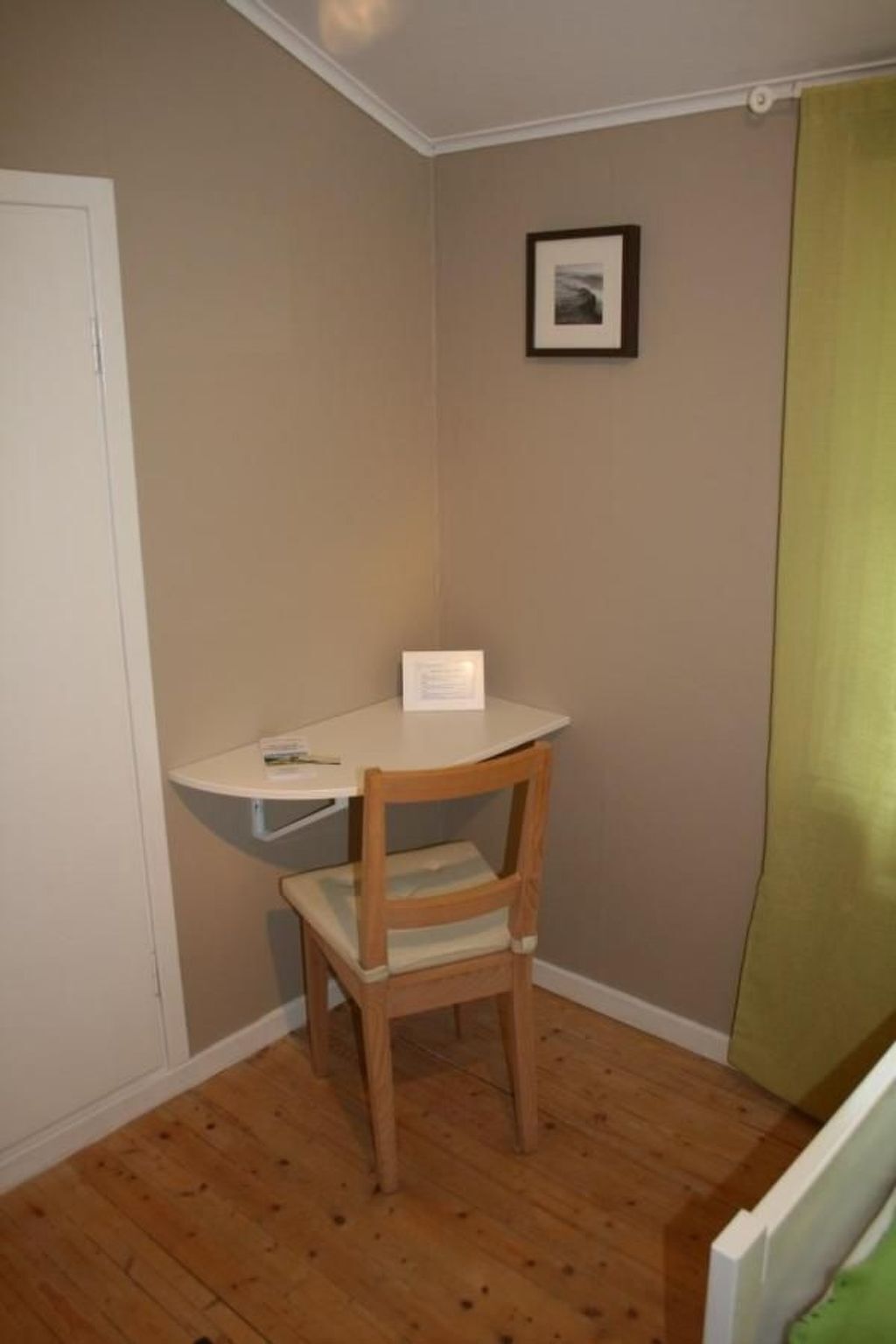 Ein Eckzimmer mit weißem Schreibtisch, einem Holzstuhl, einem gerahmten Bild an der Wand und einem grünen Vorhang.