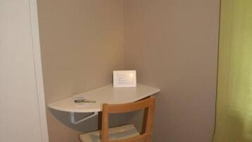 Ein Eckzimmer mit weißem Schreibtisch, einem Holzstuhl, einem gerahmten Bild an der Wand und einem grünen Vorhang.
