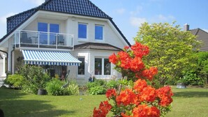 Ein zweistöckiges Haus mit Balkon, einem Garten mit roten Blumen und einem blau-weiß gestreiften Markisen.