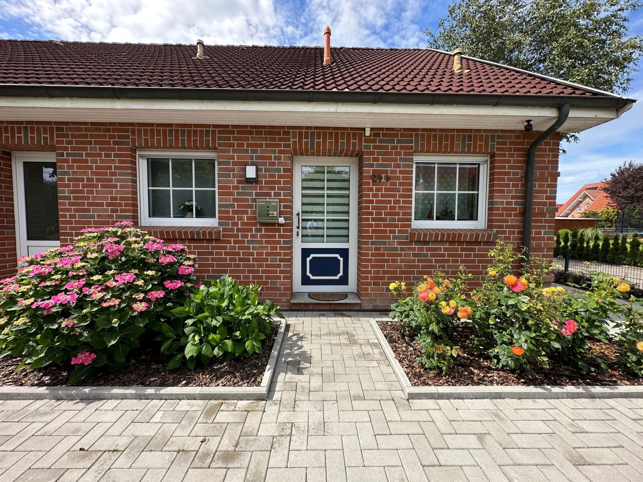 A two-story brick building with a red-tiled roof, a front door, two windows, and a small porch area with potted plants.