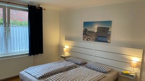 Ein Doppelbett mit karierter Bettwäsche, Nachttischlampen, ein Fenster mit Vorhängen und ein Wandbild mit Strandmotiv.