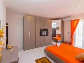 A modern bedroom with a large wardrobe, a bed with orange bedding, a TV on a shelf, and a small table with decorative items. - Schöne, strandnahe Ferienwohnung in San Tumas by Interhome (Marsascala)