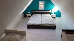 Une chambre avec un plafond haut, un lit avec une literie à motifs, une fenêtre et une table de chevet avec une lampe.