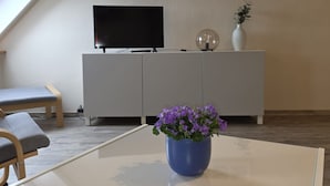 Ein Wohnzimmer mit Fernseher auf einem weißen Schrank, einer blauen Vase mit lila Blumen auf einem Tisch und einem gerahmten Bild an der Wand.