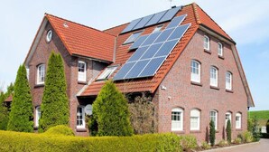 Ein zweistöckiges Backsteinhäuschen mit Solaranlage auf dem Dach, umgeben von gepflegten Hecken und Bäumen.