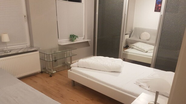 Un dormitorio moderno con una cama individual, una mesita de noche, un radiador, una ventana con persianas y un estante pequeño con una planta.