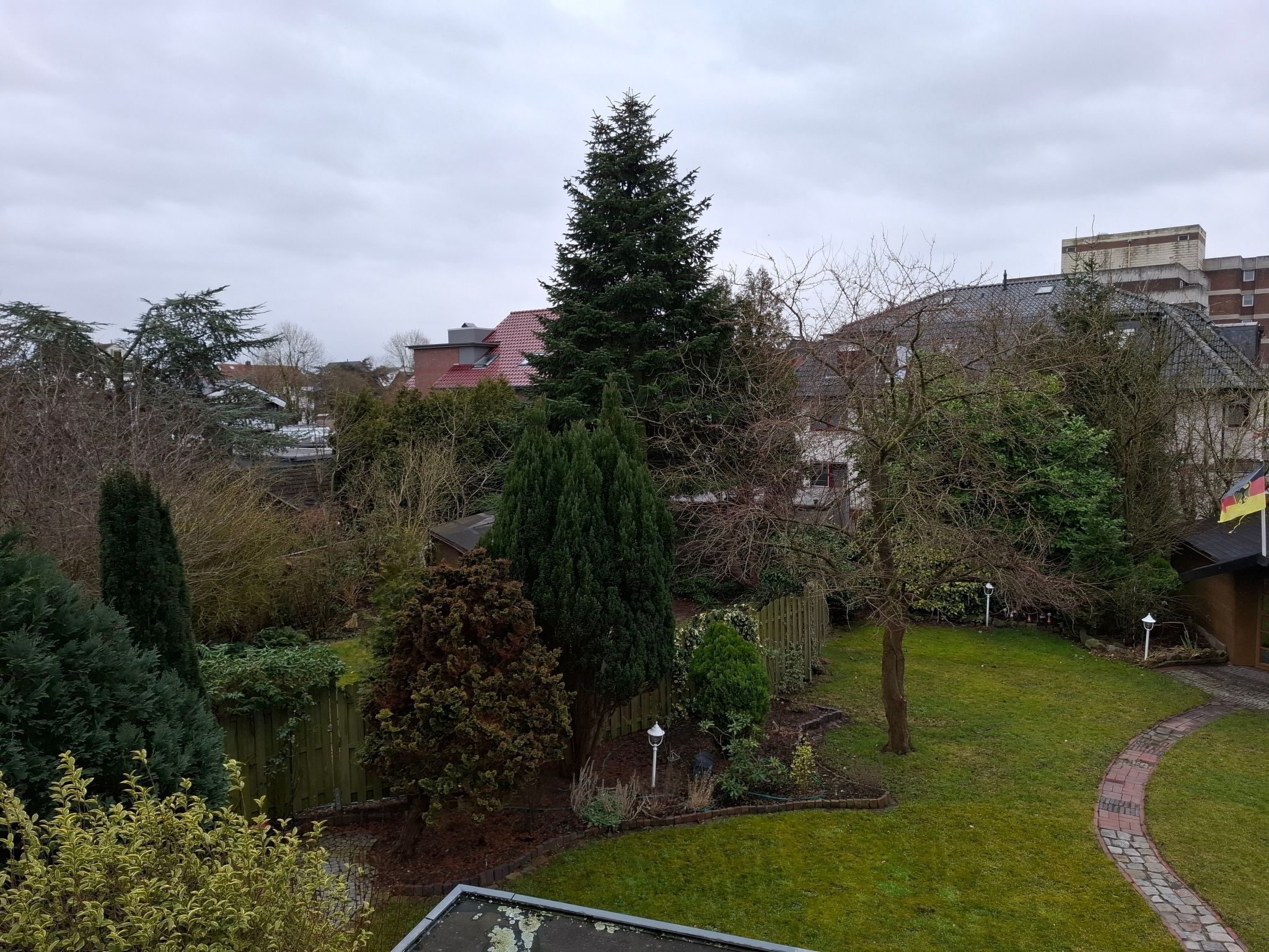 A residential garden with a variety of trees and shrubs, a paved pathway, and a small lawn area.