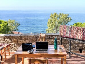 A table with a laptop, headphones, a tablet, a green water bottle, and a cup, set against a stone wall with a view of the sea and trees. - Blue Elefant by Interhome (Agios Nikolaos)