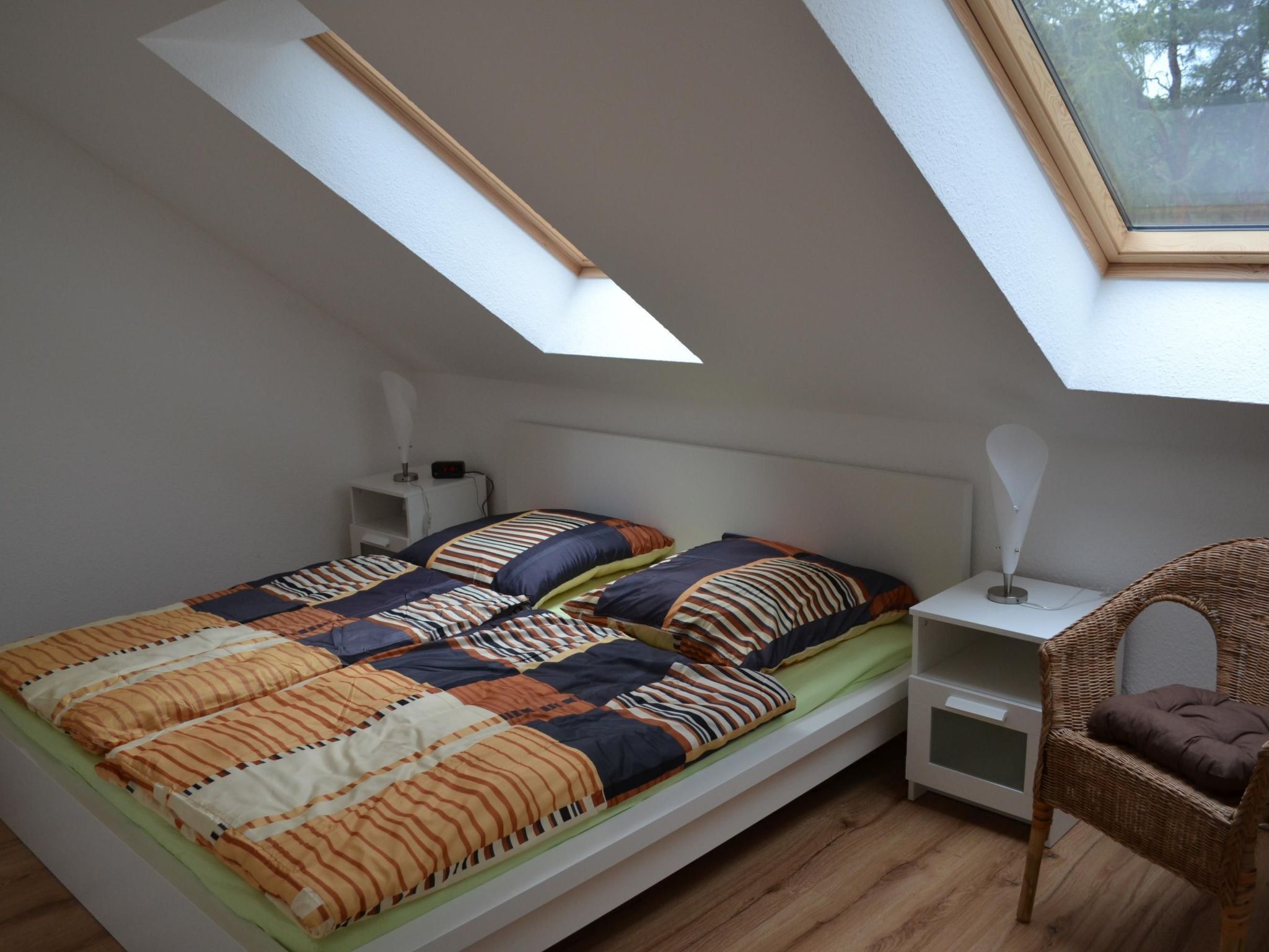 A small, single-bed room with a sloped ceiling and skylight, a bedside table with a lamp, and a wicker chair.