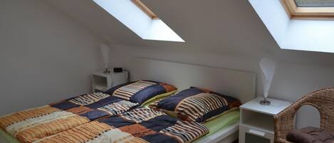 A small, single-bed room with a sloped ceiling and skylight, a bedside table with a lamp, and a wicker chair.