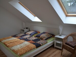 Ein kleines Zimmer mit einem Einzelbett, geneigter Decke und Dachfenster, einem Nachttisch mit Lampe sowie einem Korbstuhl.