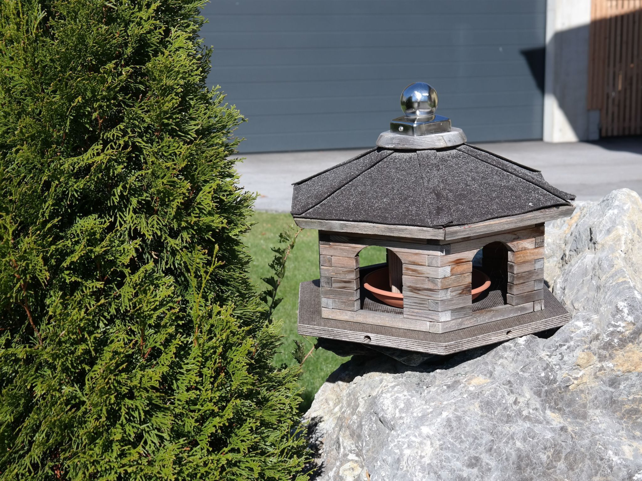 A birdhouse with a metal top and stone base, placed on a rock.