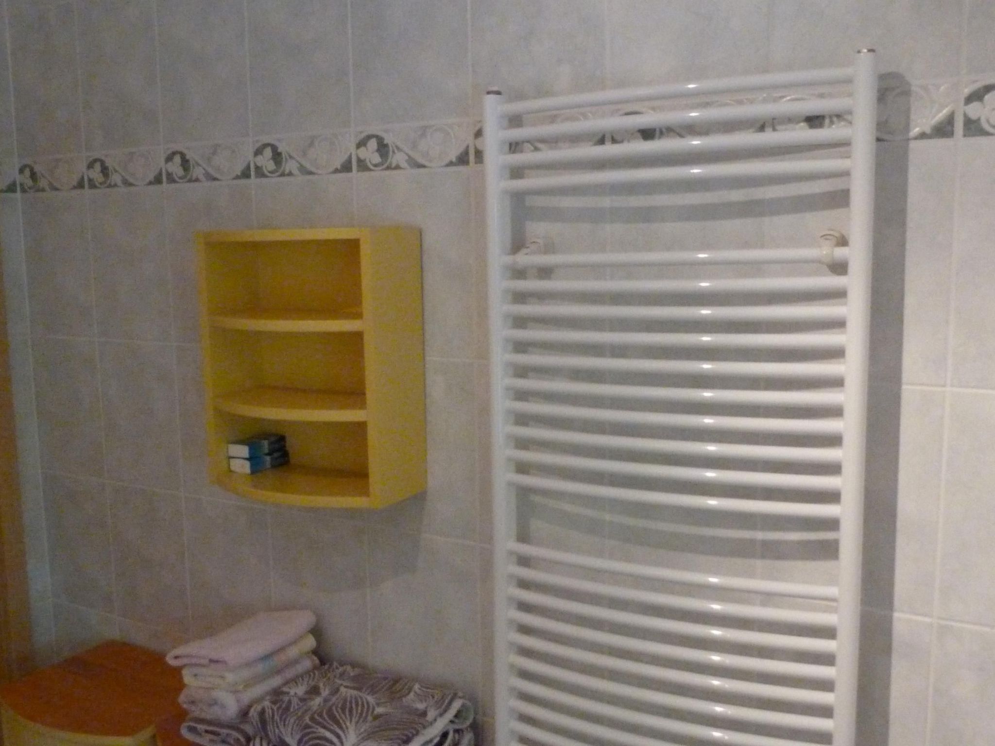 A bathroom with a white towel rack, a yellow shelf with toiletries, and a patterned towel on the floor.