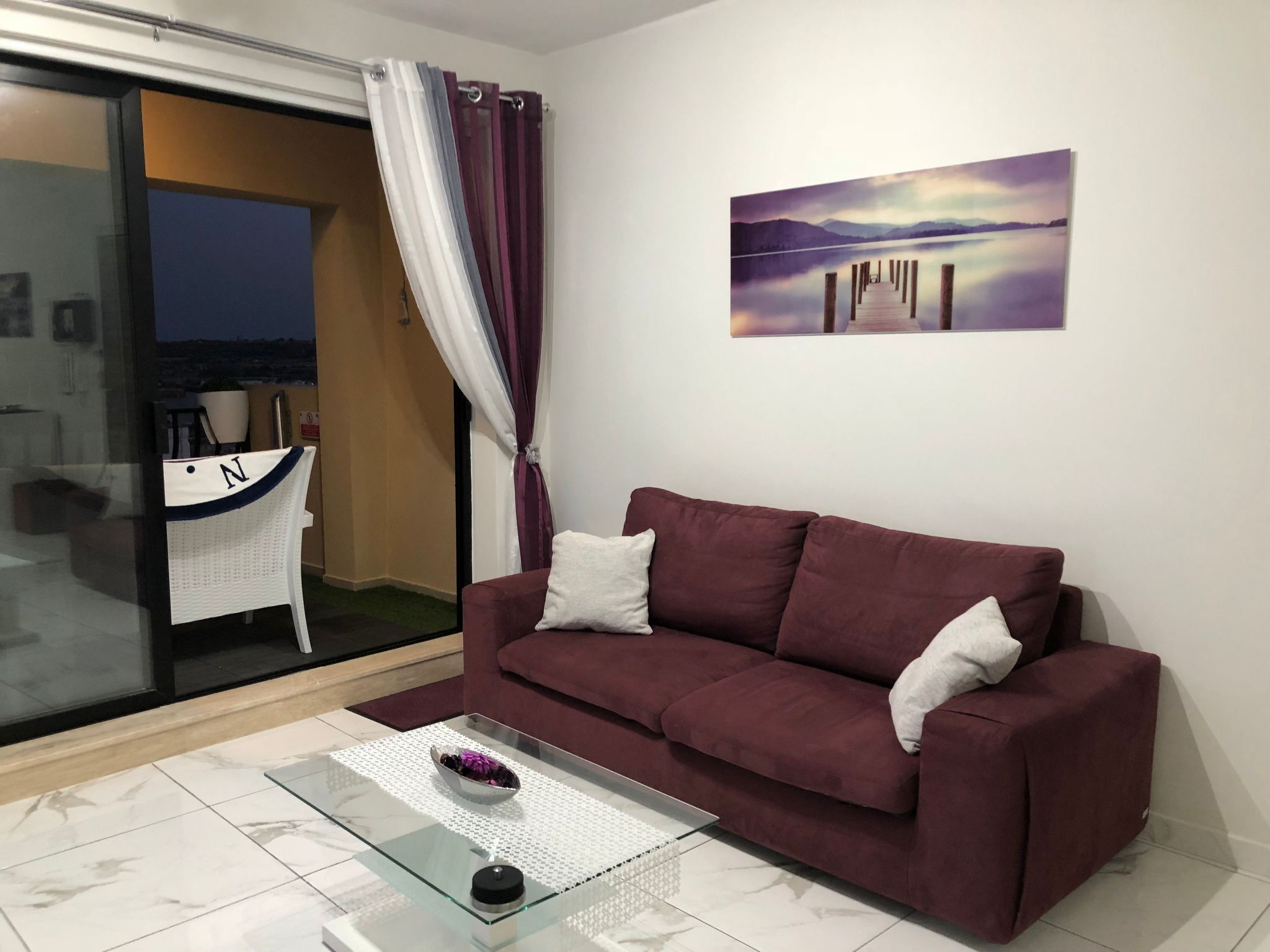 Un salon moderne avec un canapé bordeaux, une table basse en verre et un tableau représentant un lac.