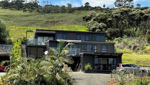 A modern two-story house with a gravel driveway, surrounded by lush greenery and a hilly landscape.