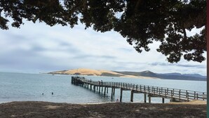 Een strand met een houten pier die zich uitstrekt over rustig water, omringd door heuvels en bomen.