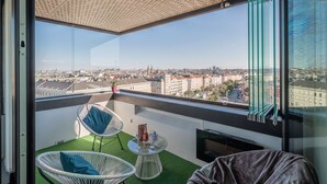Un balcón con vista a la ciudad, dos sillas de mimbre, una pequeña mesa redonda con una botella y un vaso, y un suelo de césped artificial verde.