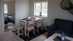 Huoneessa on puinen pöytä ja tuolit, sininen sohva, kuviollinen päiväpeiteellä varustettu sänky ja ikkuna, josta avautuu näkymä vehreään maisemaan.