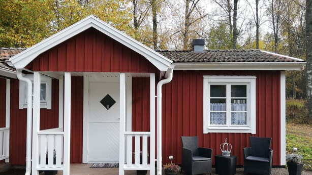 Una casa de madera roja con un porche blanco, una ventana pequeña y dos sillas de mimbre afuera.