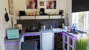 Cocina compacta con horno integrado, cafetera y lavabo. Hay objetos decorativos en los estantes y una ventana con vistas a zonas verdes.