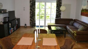 Un salon comprenant une table à manger dressée pour deux personnes, un canapé, un téléviseur posé sur un meuble et une fenêtre avec des rideaux.