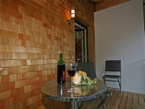 Ein Glastisch mit einer Flasche, einem Weinglas, einer Kerze und Sonnenblumen auf einem Balkon mit einer Ziegelwand und einem Stuhl.
