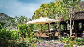 Un espace de restauration en plein air, avec une table et des chaises placées sous une tonnelle, entouré d’un jardin agrémenté de diverses plantes et fleurs.