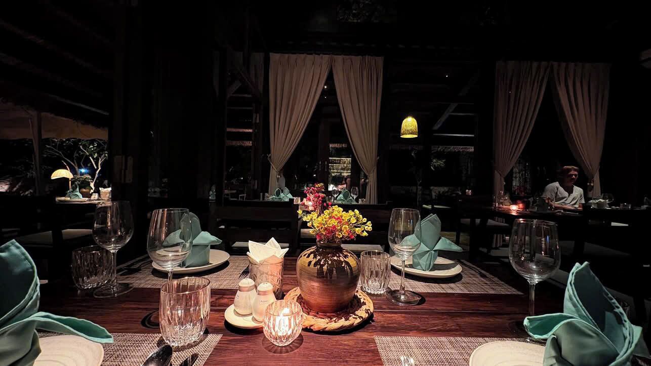 A dimly lit restaurant with a wooden table set for two, featuring a vase of flowers, glasses, and a candle.