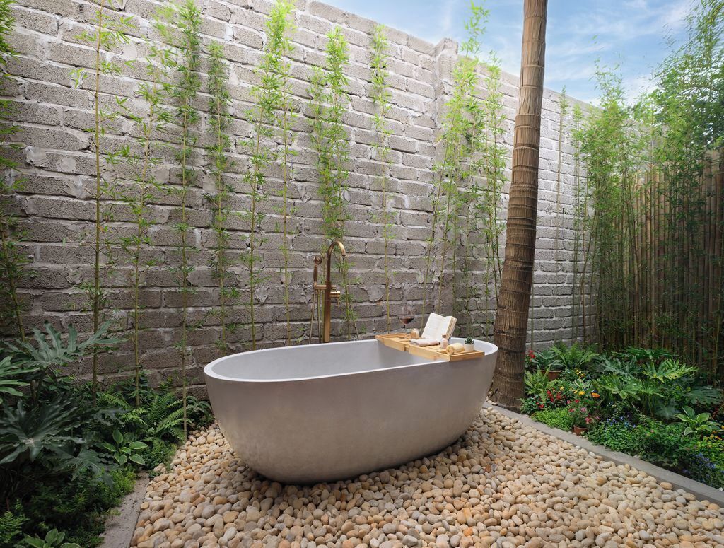 A freestanding bathtub in a garden setting with a brick wall backdrop and lush greenery.