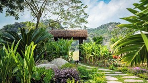 A garden pathway leading to a thatched-roof structure surrounded by lush tropical plants and a mountainous backdrop. - La Lua Resort Ninh Binh (Tay Hoa Lu)