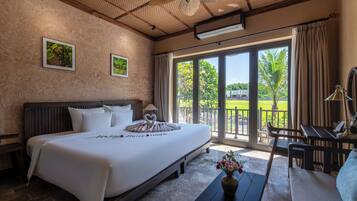 Habitación de hotel con una cama grande, vista a un jardín a través de puertas correderas y un balcón con mesa y sillas.