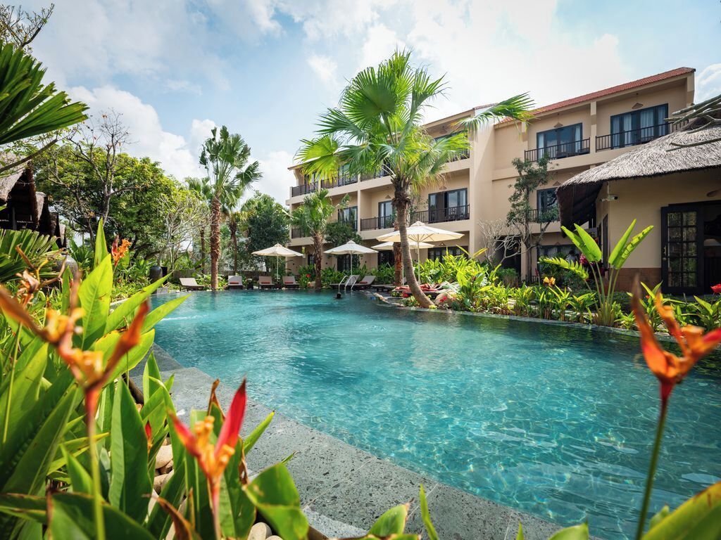 A pool area with lounge chairs and umbrellas, surrounded by tropical plants and a building with balconies.
