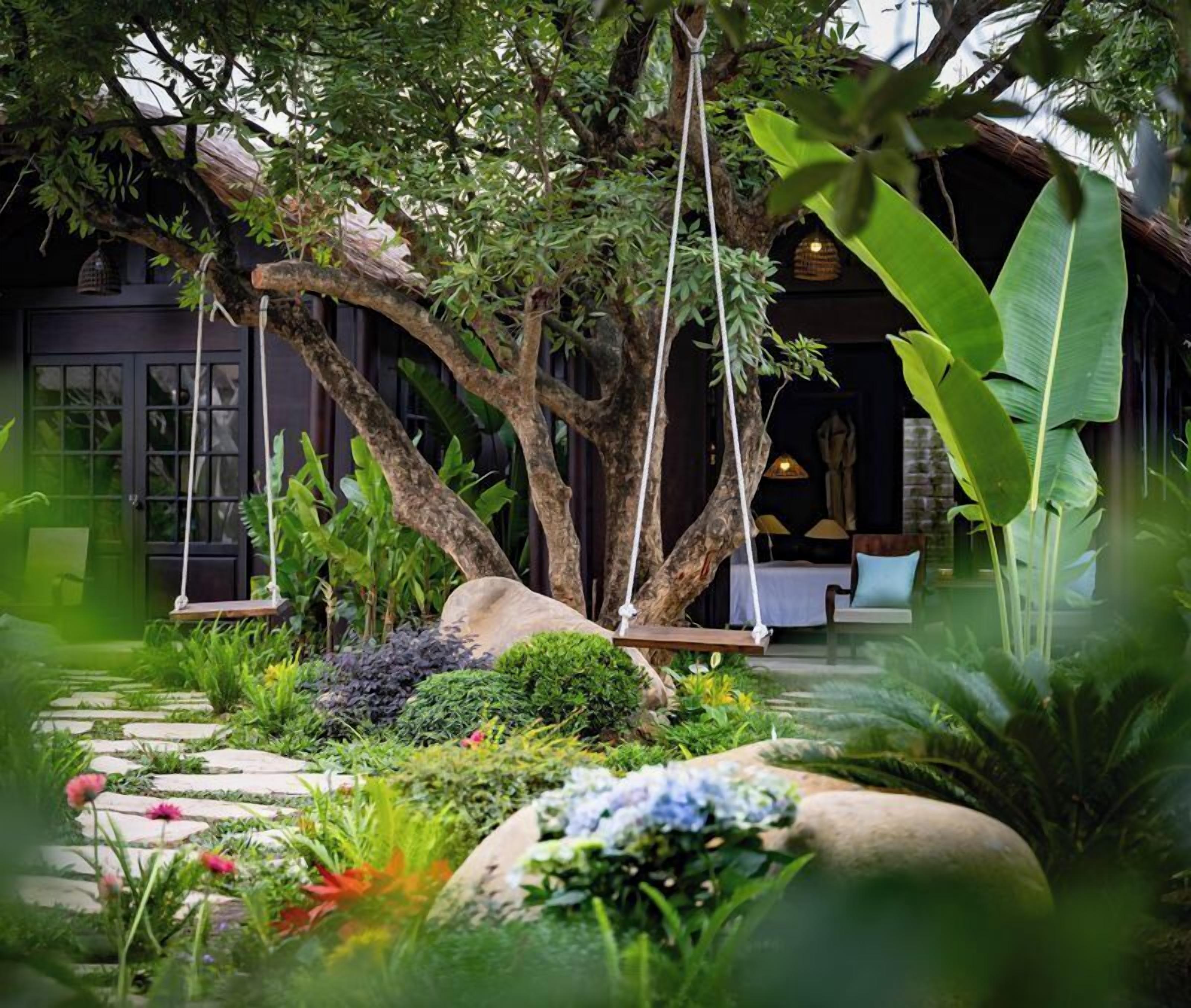 A lush garden with a swing, a stone pathway, and a building with a dark facade and large windows.