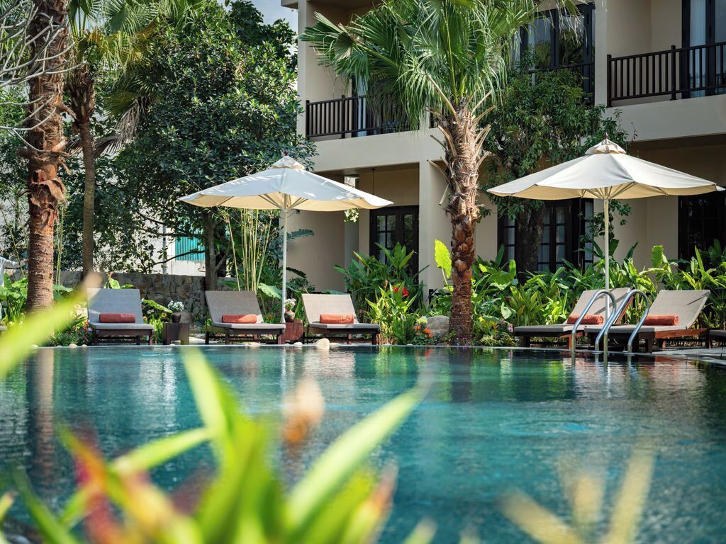 A pool area with lounge chairs and umbrellas, surrounded by lush greenery and a building with balconies.