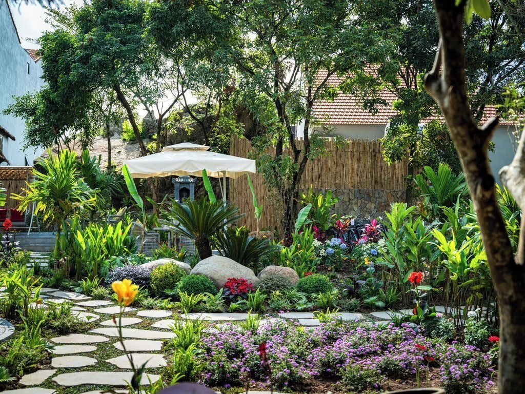 A garden with a stone pathway, various plants, a large umbrella, and a rustic fence.