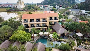 A resort with a swimming pool, thatched-roof structures, and a view of a mountain and a large building in the background. - La Lua Resort Ninh Binh (Tay Hoa Lu)