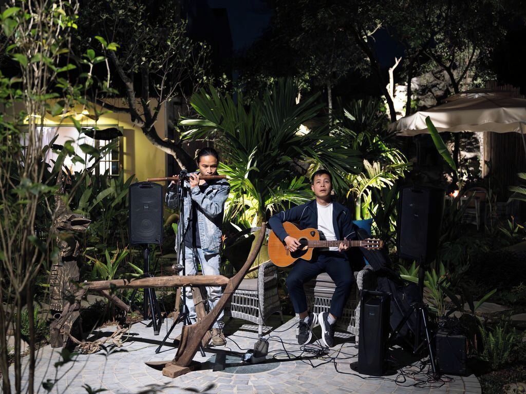 Two musicians performing outdoors with musical instruments and speakers, surrounded by greenery and a rustic setting.