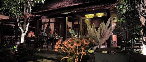 An outdoor dining area with tables and chairs, surrounded by plants and a thatched roof structure.
