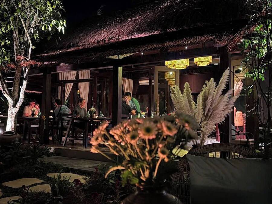 An outdoor dining area with tables and chairs, surrounded by plants and a thatched roof structure.