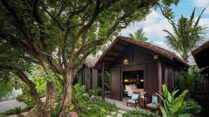 A rustic-style building with a thatched roof, surrounded by lush greenery and a stone pathway leading to a seating area with outdoor furniture. - La Lua Resort Ninh Binh (Tay Hoa Lu)