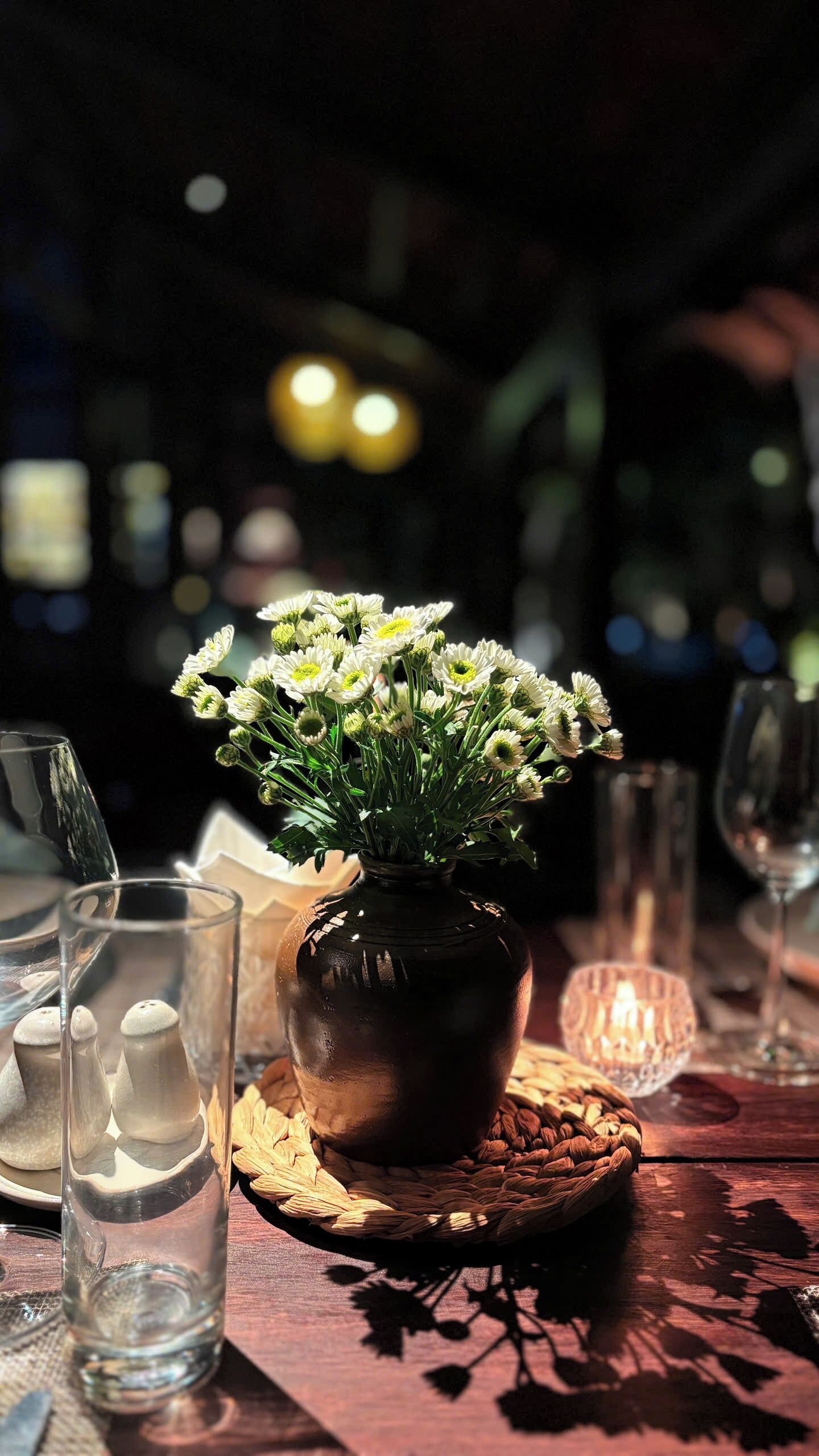 A table with a dark brown wooden surface, a clear glass vase with white flowers, salt and pepper shakers, a woven coaster, and a glass of water.