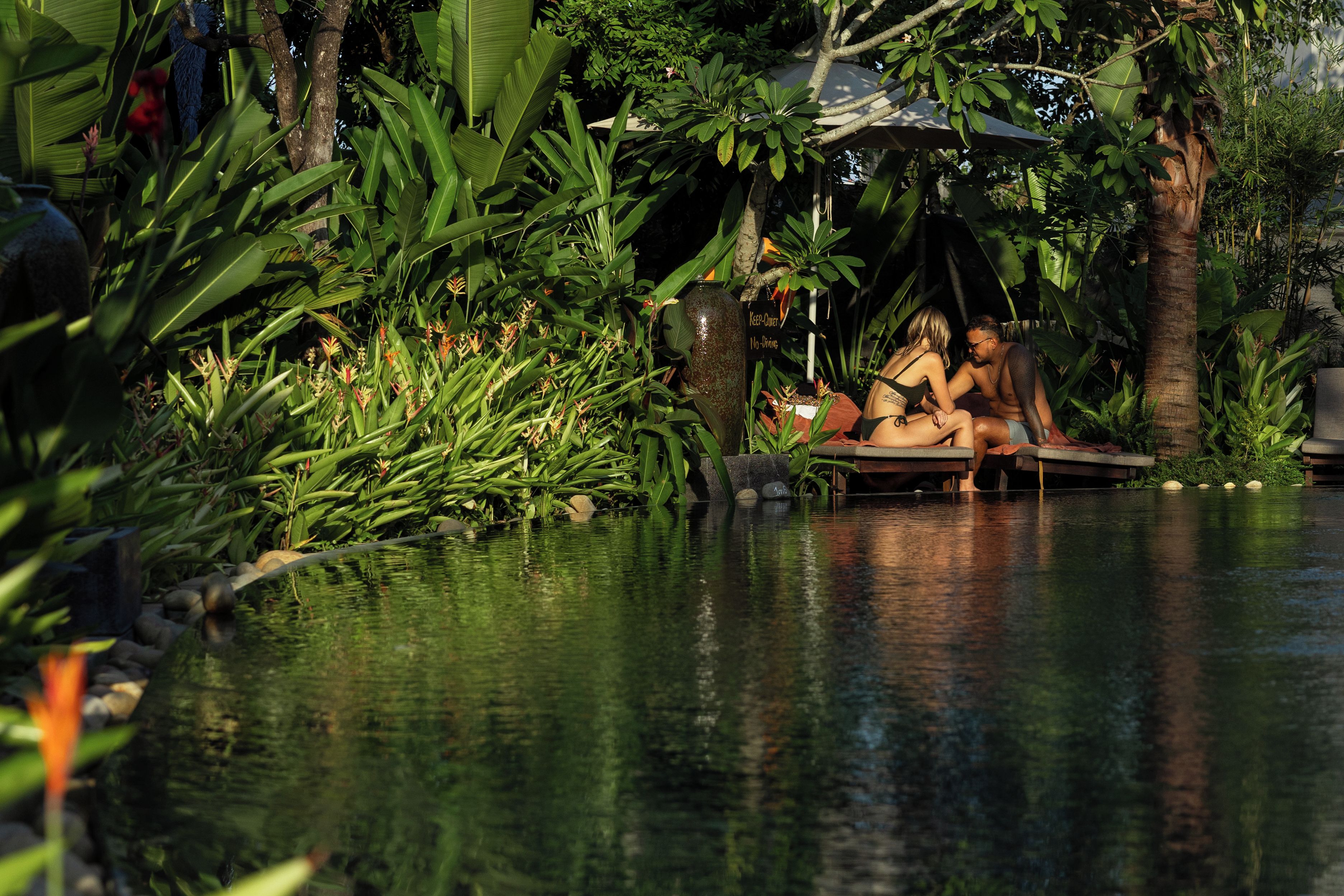 A pool area with tropical plants and a couple sitting on a bench.